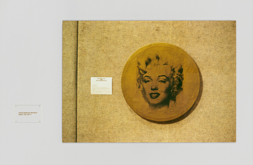 A round gold panel with a stylized blonde woman's face hangs on a textured beige wall.