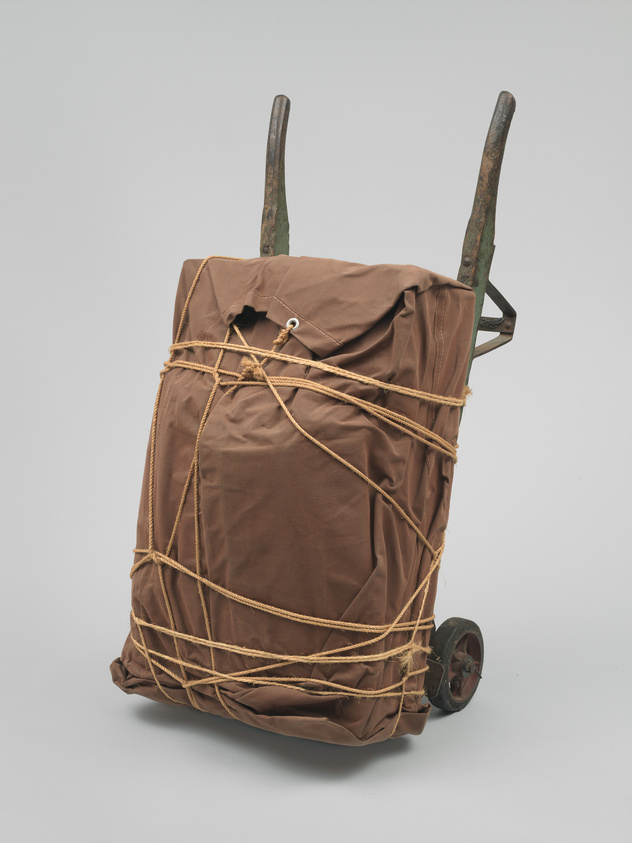 A lumpy, rectangular package wrapped in brown tarp and rope and supported by an aged, wooden hand truck.