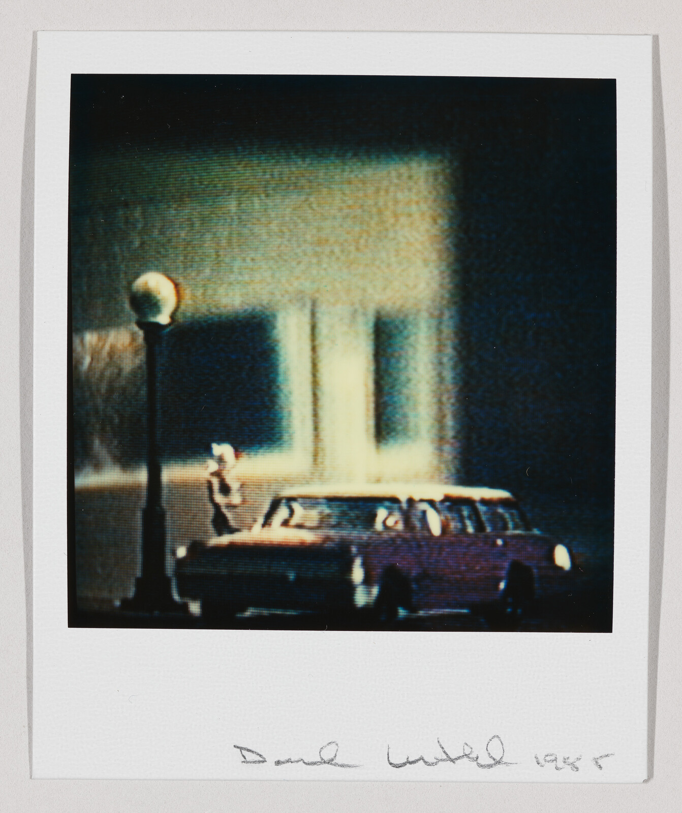 A person stands beside a parked vintage car under a streetlamp at night.