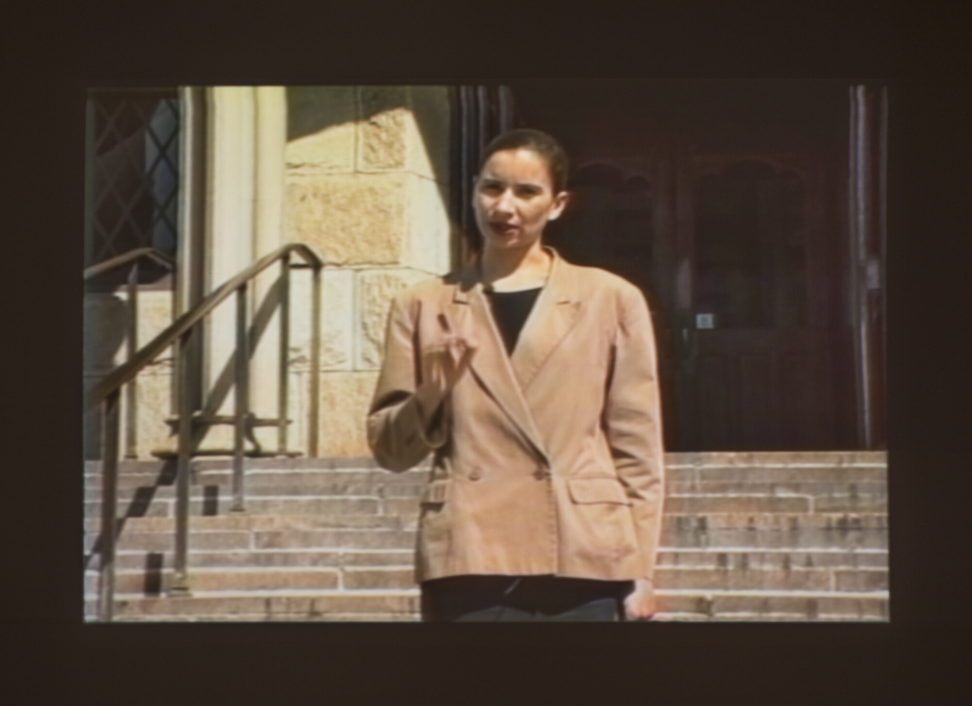 A woman in a tan blazer stands on stone steps, holding up her hand and gesturing with her fingers.