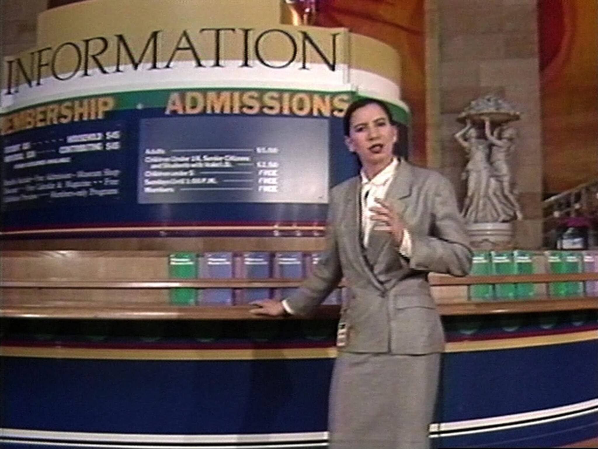 A woman in a gray suit gestures while standing at an information and admissions desk.