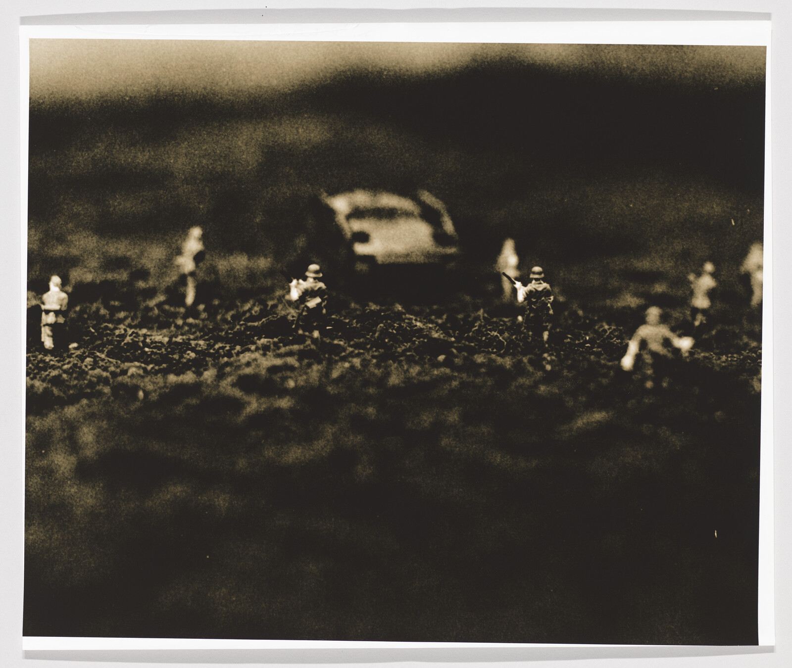 Small toy soldiers advance across a dark, grainy terrain toward a blurred vehicle in the background.