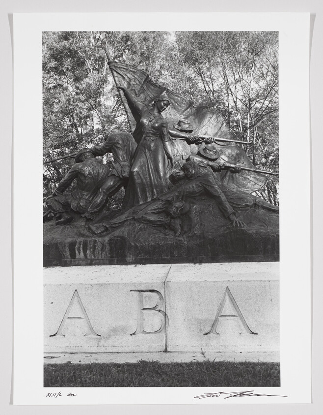 A black and white photograph of a dynamic sculpture depicting several figures in a dramatic pose, with one central figure holding a flag aloft. The base of the sculpture has the letters "ABA" inscribed on it. The image is numbered "XIII/16" and signed by the artist in the lower margin. Trees form a natural backdrop to the sculpture.