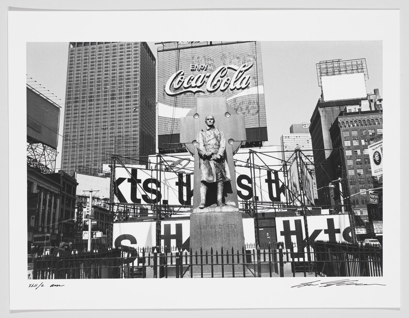 Black and white photograph of a cityscape featuring a statue in the foreground with large billboards, including a prominent Coca-Cola advertisement, in the background. Skyscrapers tower over the scene, and scaffolding with large letters is visible. The image has a white border with handwritten signatures at the bottom.