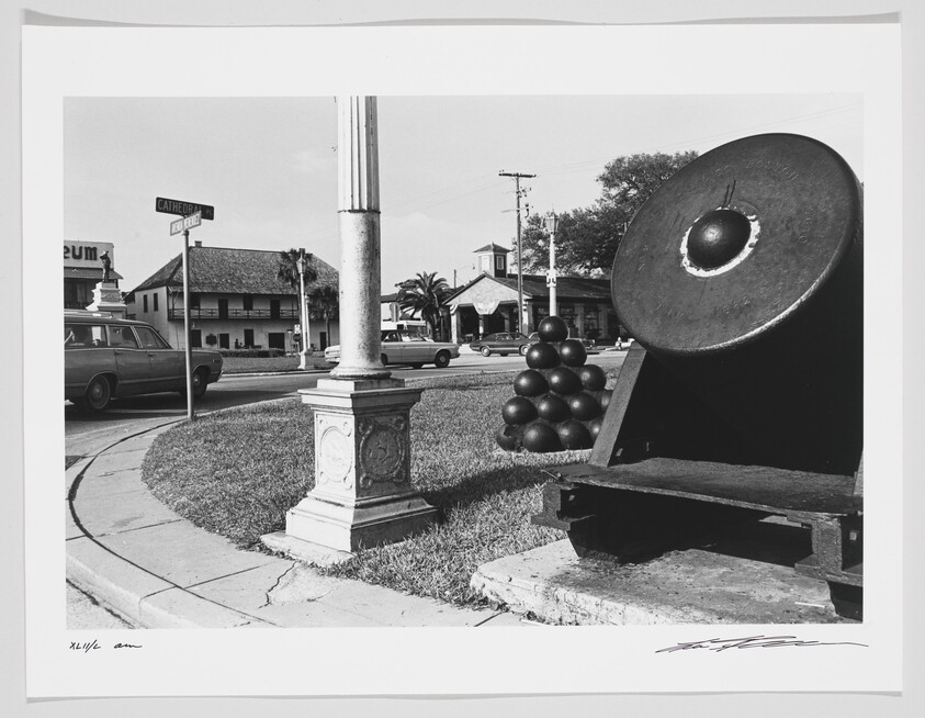 Black and white photograph depicting an outdoor scene with a large, old cannon in the foreground on the right, mounted on a display stand with a stack of cannonballs behind it. In the background, there's a street corner with a sign reading "CATHEDRAL" and a vintage station wagon parked on the side of the road. A building with the sign "MUSEUM" is visible across the street, and there are palm trees and utility poles scattered throughout the scene. The image has a border with handwritten signatures at the bottom.