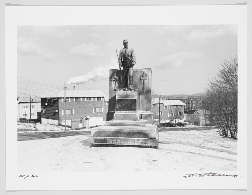 A black and white photograph depicting a statue of a standing figure on a pedestal with inscriptions, set against a backdrop of a residential area with snow-covered ground. Smoke rises in the distance from a chimney, and hills are faintly visible on the horizon. The image includes a handwritten signature and date in the lower right corner.