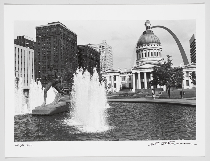 A black and white photograph capturing an urban scene with a statue in the foreground, surrounded by water fountains. In the background, the iconic Gateway Arch looms beside a domed building, possibly a capitol or courthouse, with several other high-rise buildings. There is a pedestrian walking on the sidewalk, and the image has signatures at the bottom.