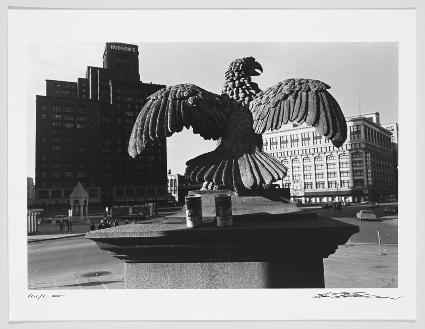 A black and white photograph featuring a close-up of a sculpted eagle with outstretched wings, perched atop a pedestal. In the background, a large building with the sign "HUDSON'S" on its top is visible, along with other urban structures. On the pedestal next to the eagle sculpture, there are two cans, one of which is labeled "Faygo Grape." The foreground is dominated by the eagle's detailed feathers, while the urban landscape provides context in the distance. The image also includes the artist's signature in the lower right corner.