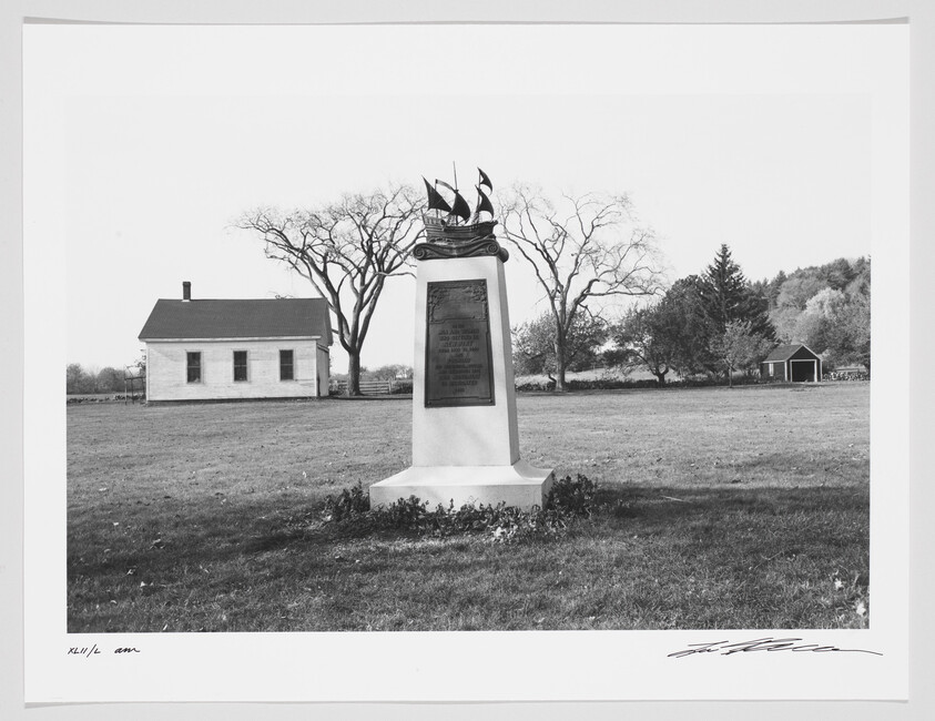 A black and white photograph depicting a historical monument with an inscription plaque and a sculpture of an eagle with spread wings on top. In the background, there's a small white house with a chimney to the left and a small barn to the right, surrounded by a grassy field with scattered trees. The image has a handwritten signature at the bottom right corner.