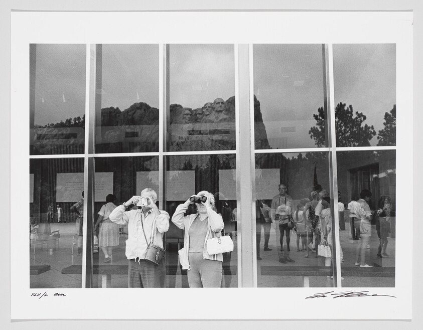 A black and white photograph capturing the reflection of Mount Rushmore in a large window, with two visitors in the foreground looking up through binoculars. The reflection merges with the interior scene, showing other visitors milling about, creating a layered visual effect. The image is signed by the photographer at the bottom.