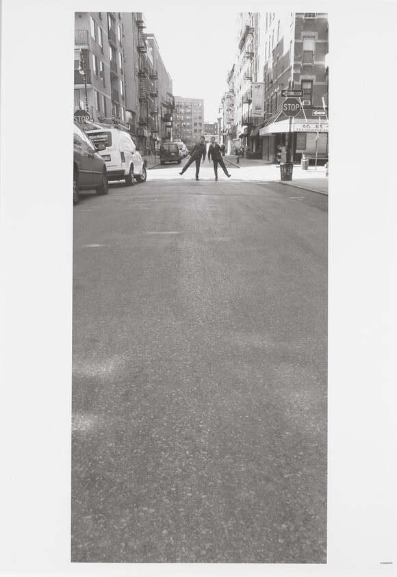 A black and white photograph capturing two individuals standing in the middle of a city street, arms outstretched towards each other, creating a mirrored pose. The street is lined with parked cars and buildings, and there's a stop sign and one-way sign visible on the right. The scene is devoid of traffic, giving a sense of stillness to the urban environment.