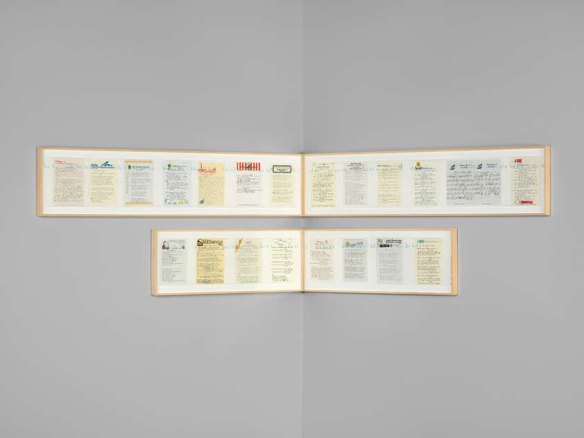 A long framed display containing multiple pages of handwritten letters and documents, mounted on a light gray wall.