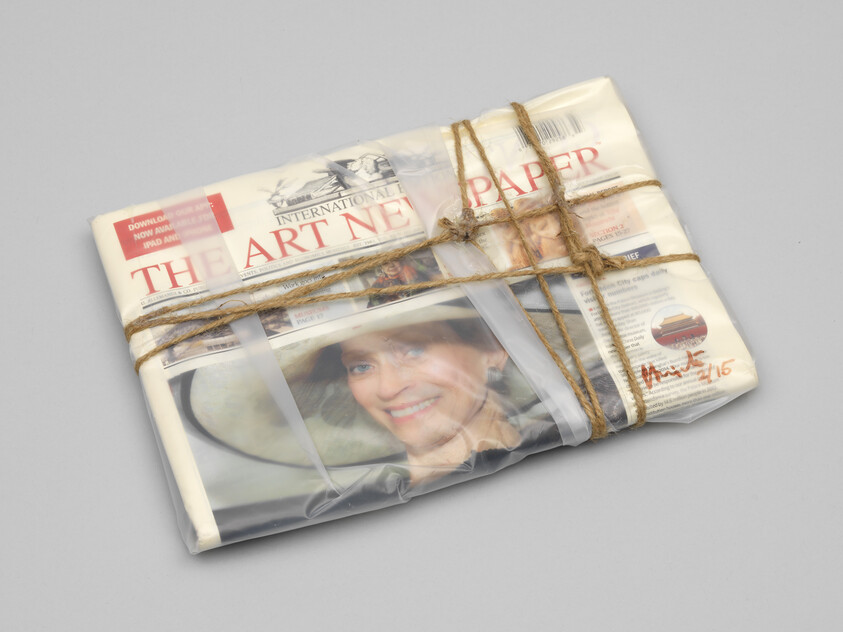 A bundle of newspapers titled "THE ART NEWSPAPER" tied with twine and partially wrapped in plastic, with visible headlines and a woman's face on the front page. There's a handwritten note on the plastic wrap that appears to be a signature with the numbers 4/15.