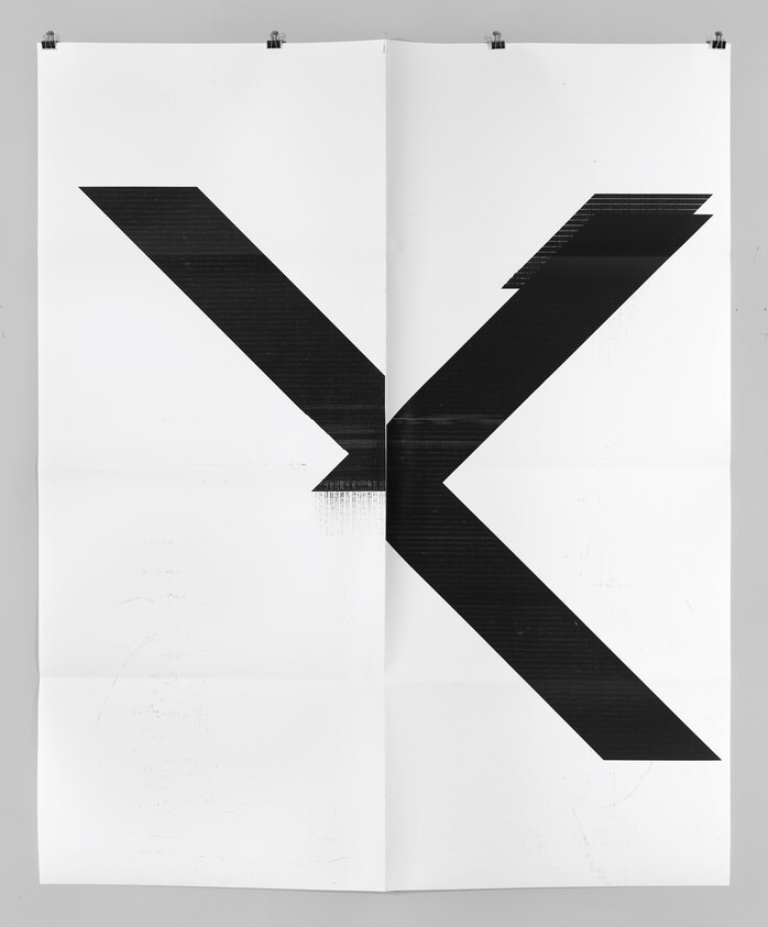 Large black angular K-shaped graphic on a folded white poster clipped to a wall.