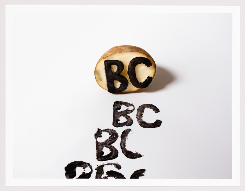 A potato cut in half with the letters "BC" carved into it, sitting above several black ink prints of the same letters on a white background.