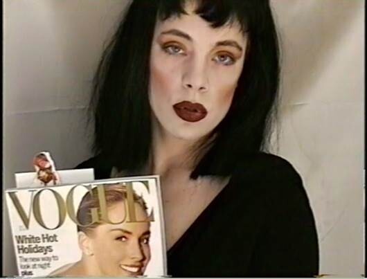 A woman with dark hair holds up a Vogue magazine showing a smiling model on the cover.