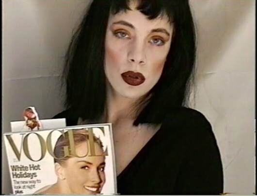 Person with dark hair and bold makeup holds a Vogue magazine toward the camera.