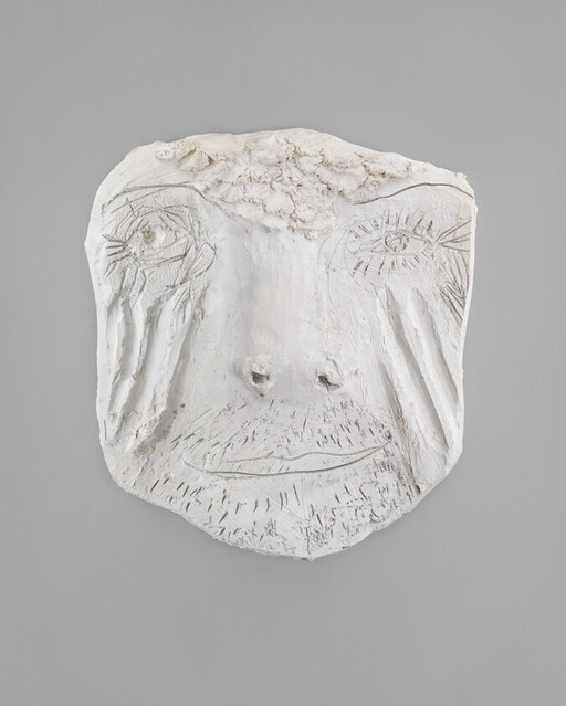 A white plaster relief face with carved eyes, nose, mouth, and textured forehead hangs on a gray wall.
