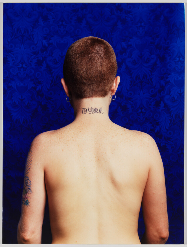 Person with shaved head and two ear hoops showing a small tattoo on the back of the neck.