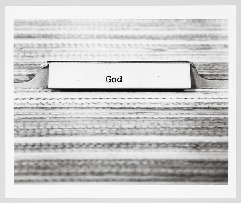 A metal file folder tab on a wood surface labeled "God."