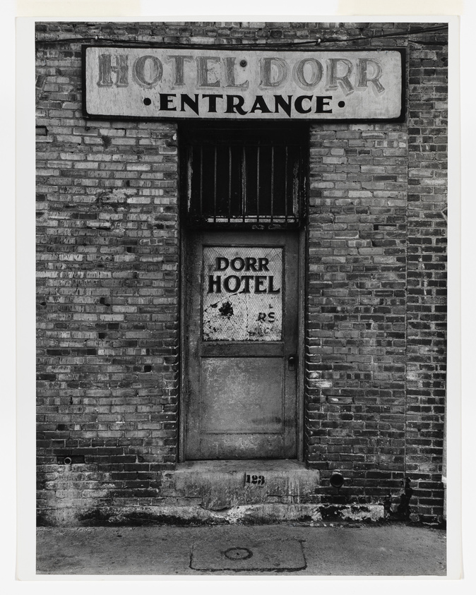 A black and white photograph of a worn and aged entrance to the Dorr Hotel, featuring a faded sign above a weathered door with peeling paint, set in a brick wall. The word "ENTRANCE" is visible below the hotel name on the sign, and the address number 129 is painted on the curb in front of the single step leading to the door.