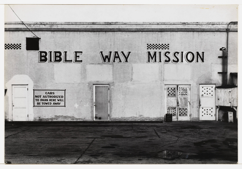 A black and white photograph of a building with "BIBLE WAY MISSION" painted in large letters on its facade. Below the text, a sign reads "CARS NOT AUTHORIZED TO PARK HERE WILL BE TOWED AWAY." The building has multiple doors and windows, some with decorative patterns, and there are checkered patterns above some windows. The ground in front of the building is wet, reflecting parts of the building.