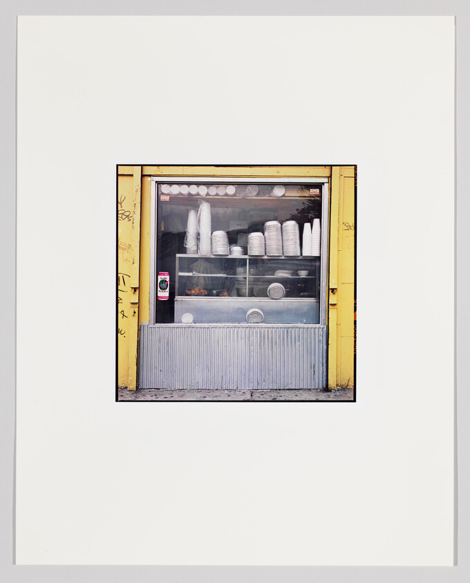 Small takeout window framed in yellow displaying stacked disposable cups and plates on shelves.