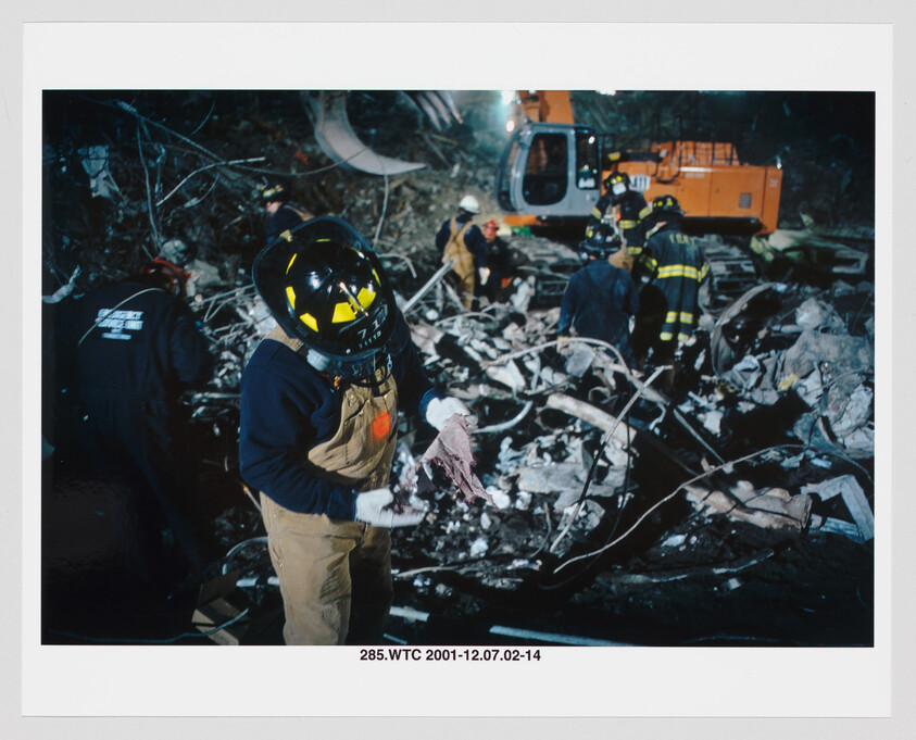 A firefighter wearing a helmet and gloves examines a piece of debris amid wreckage.