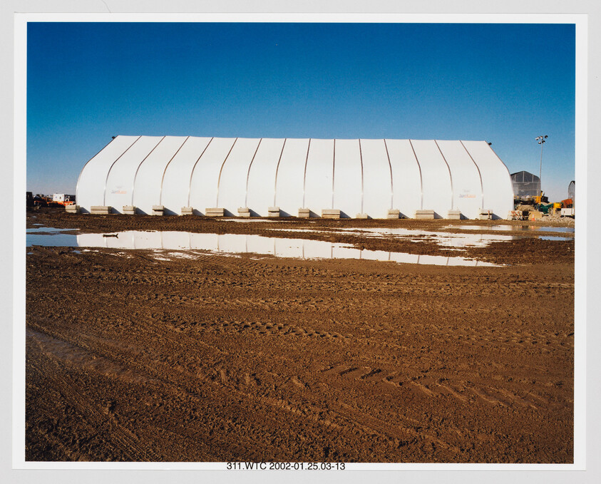 A long white temporary building stands on a muddy construction site with puddles reflecting its shape.
