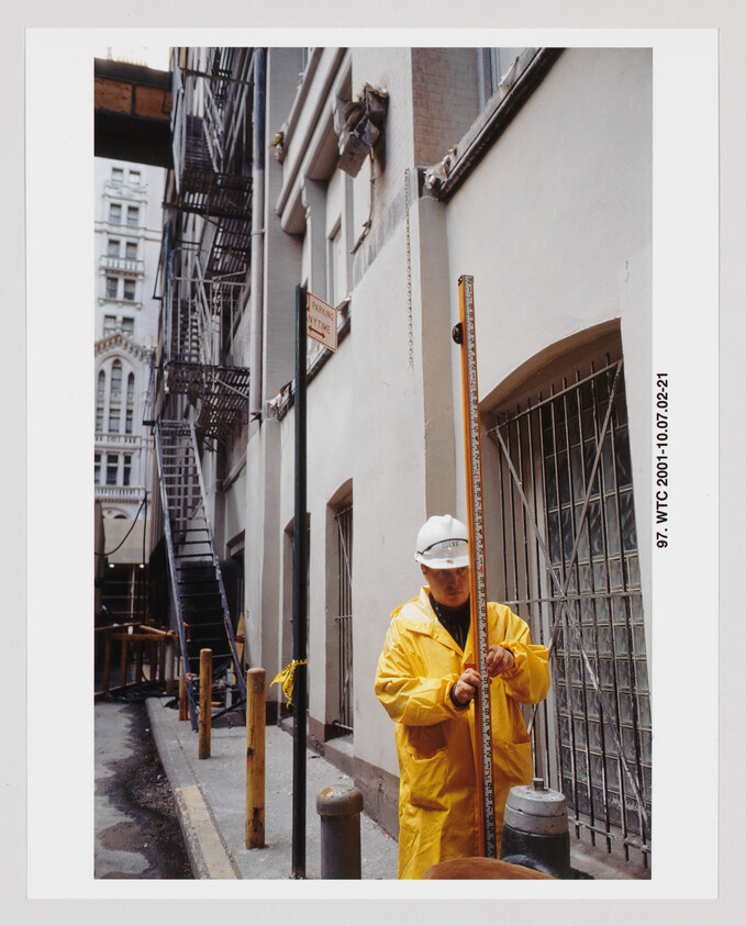 Construction worker in a yellow raincoat and hard hat measures with a leveling rod beside a building.