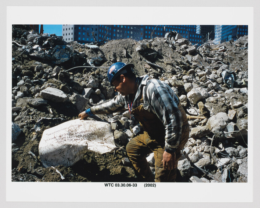 Construction worker in a hard hat points at a broken memorial stone amid rubble from a collapsed building.
