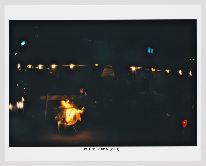 Person standing by a small open fire pit at night under a row of string lights.