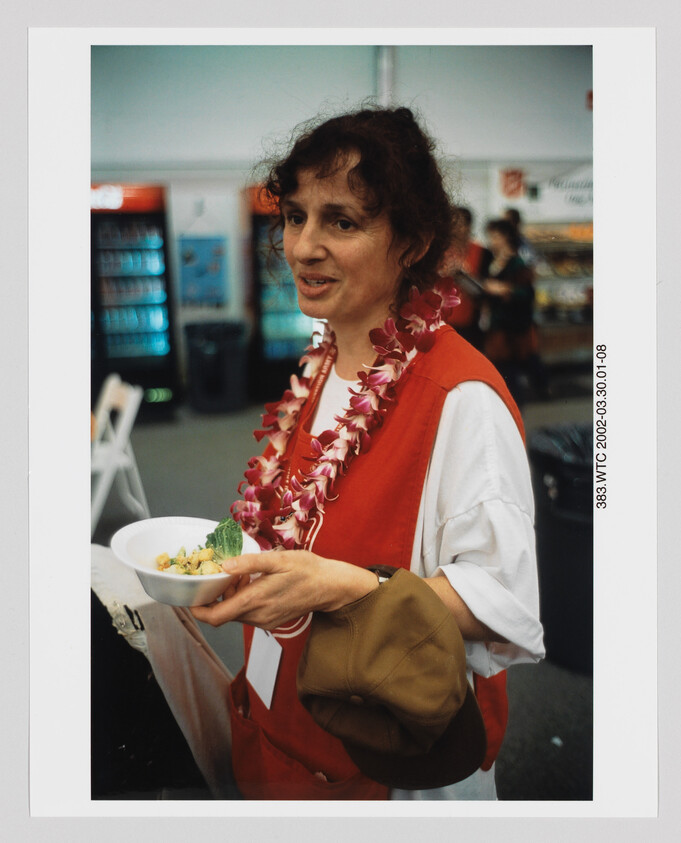 Woman wearing a flower lei and red vest holds a bowl of food and a brown cap.