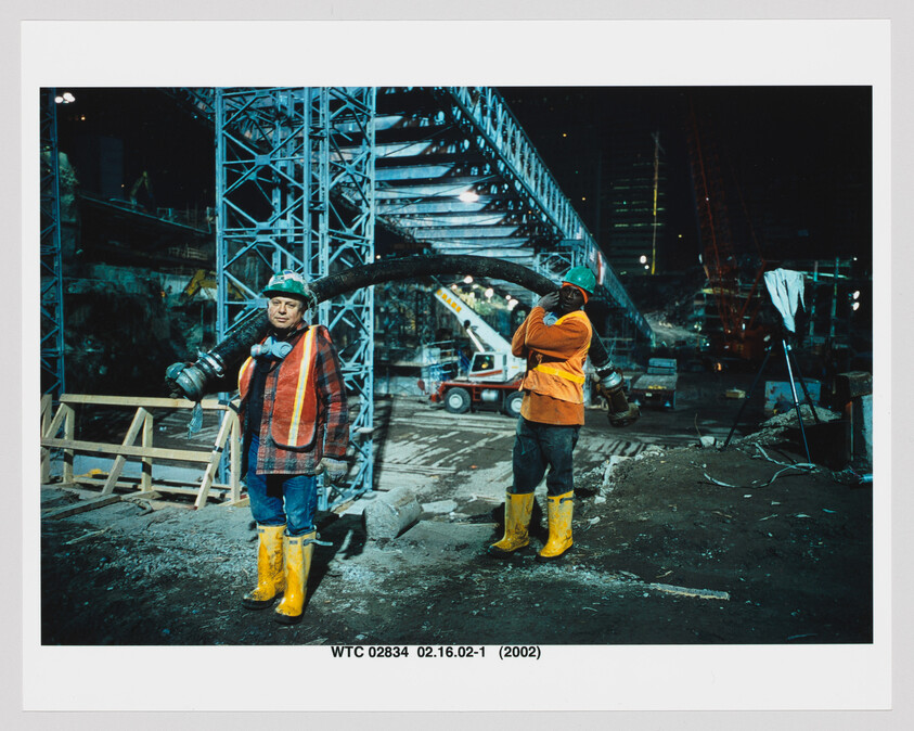 Two construction workers in safety gear carry a heavy hose at a nighttime construction site.
