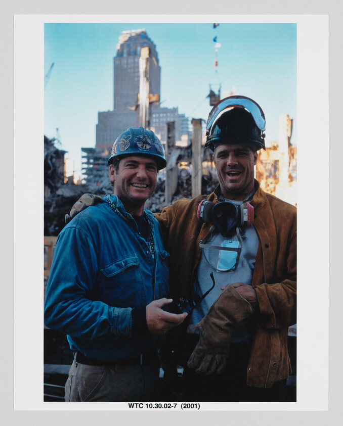 Two construction workers wearing helmets and protective gear smile at a damaged downtown construction site.
