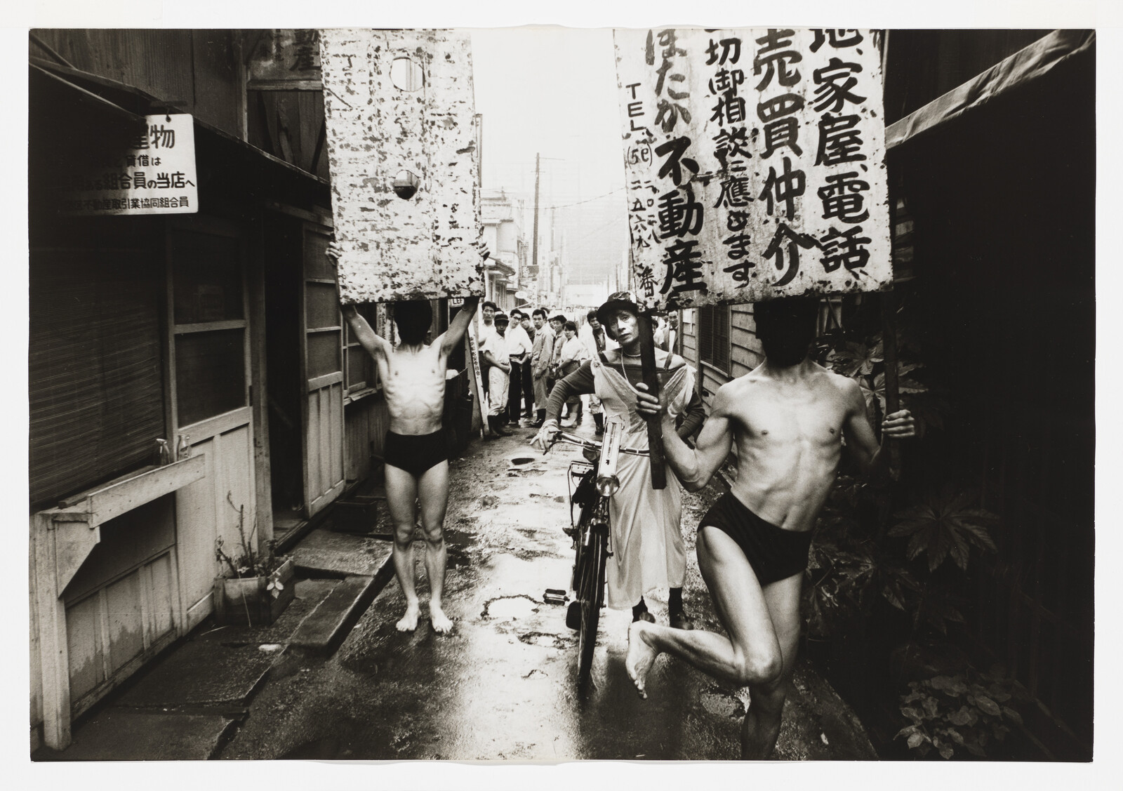 Two men in briefs carry large signs while a woman with a bicycle walks through a narrow street.