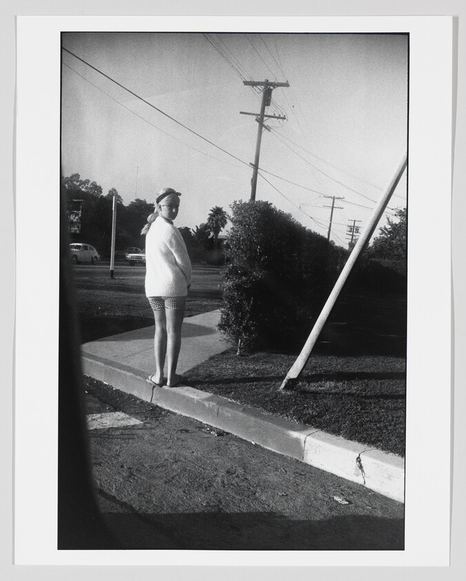 A young woman stands on a curb looking back beside bushes and a leaning utility pole.