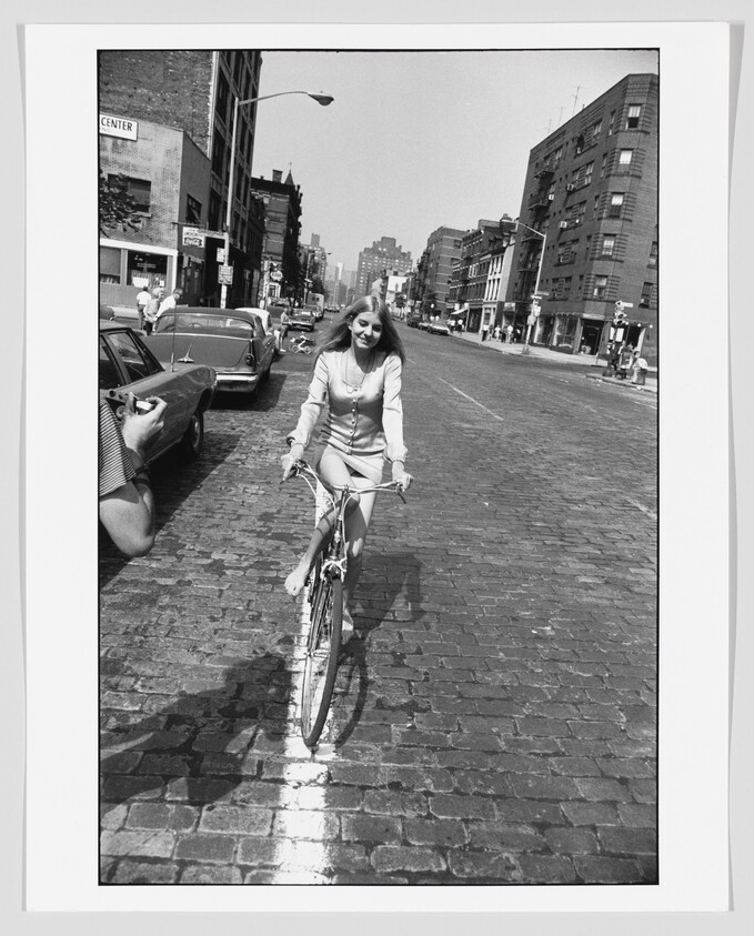 Young woman rides a bicycle down a cobblestone city street, smiling toward the camera.