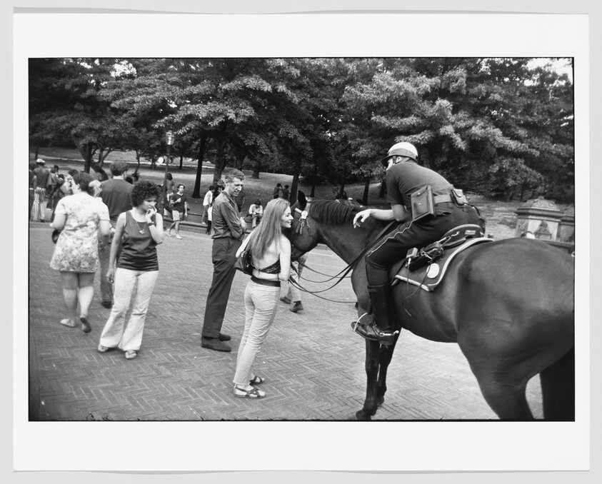 A young woman talks with a mounted police officer while other people walk in a park.