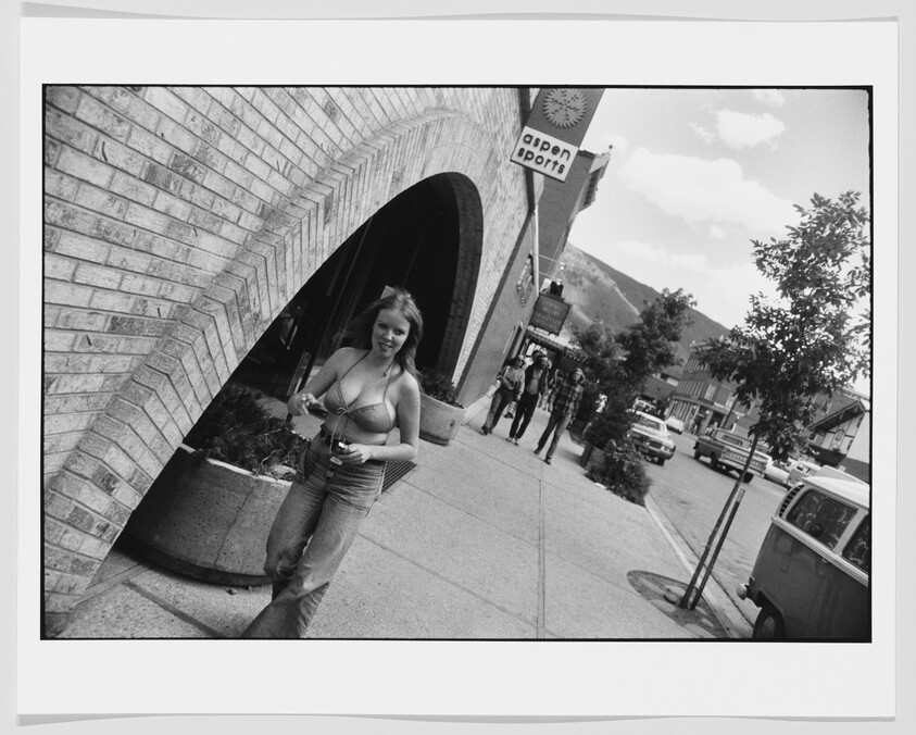 A young woman in a halter top walks along a downtown sidewalk past an arch and an Aspen Sports sign.