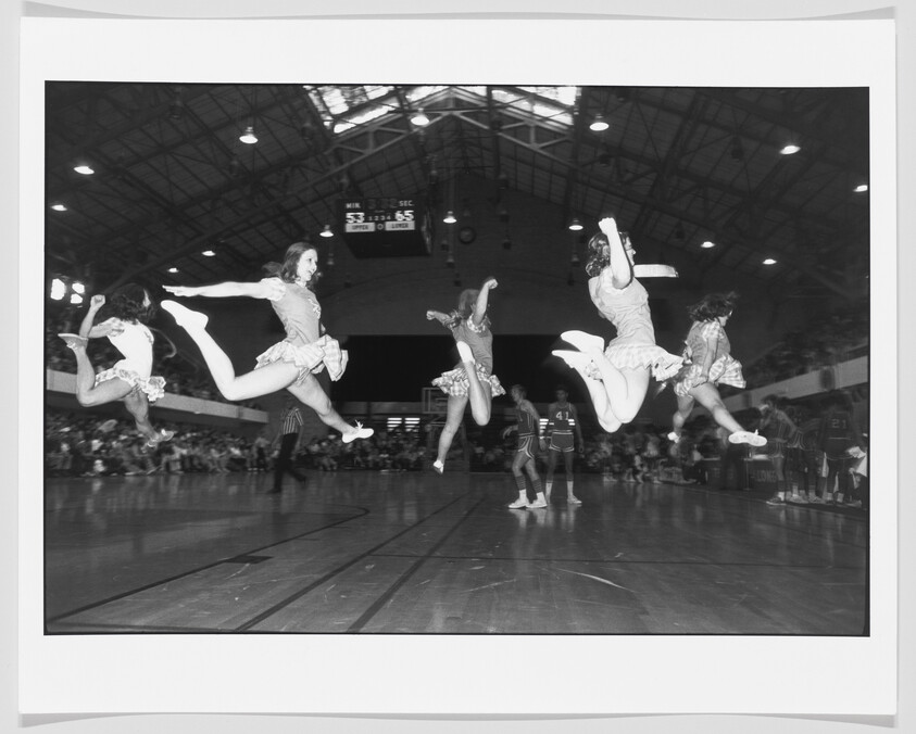 Five cheerleaders leap in synchronized jumps across a basketball court during a game.