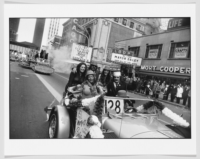A group of smiling people ride a decorated convertible car numbered 28 in a downtown parade.