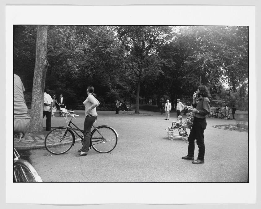 A woman stands with her bicycle hands on hips while people walk and push strollers in a park.