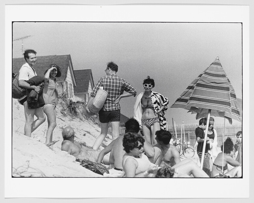 A group of beachgoers relax on a sandy shore while a woman in striped swimsuit stands near a striped umbrella.