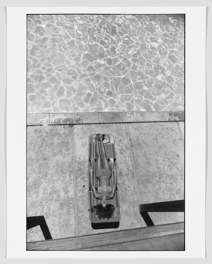 Black and white photo of a person lying on a lounge chair by the edge of a swimming pool, with the water's surface reflecting sunlight patterns. The pool's depth marker reads 5 feet.