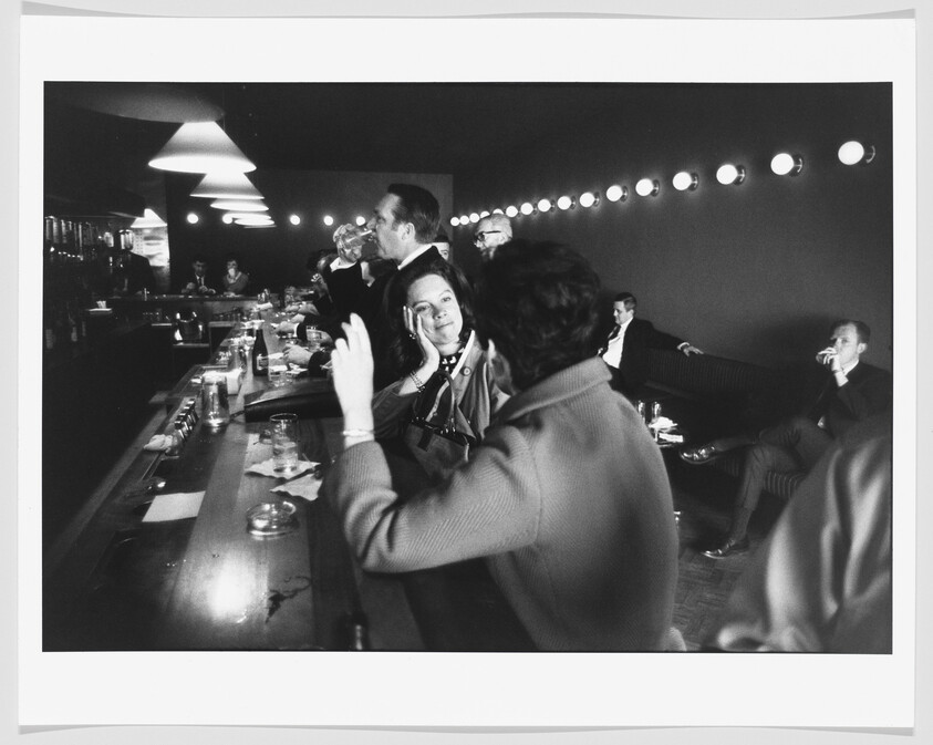 A woman at a crowded bar smiles while talking with a companion as others drink nearby.