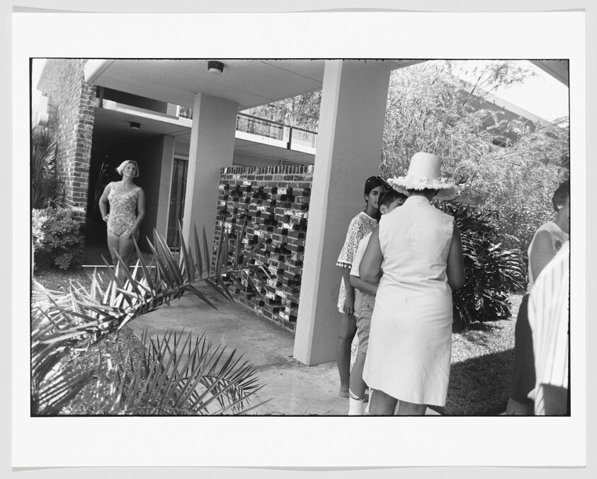 Woman in a swimsuit stands by a building entrance while a woman in a sun hat talks to children nearby.