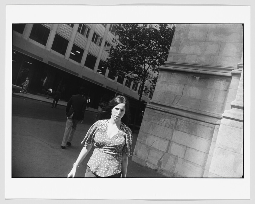A woman walks toward the camera on a city sidewalk beside a large stone building.