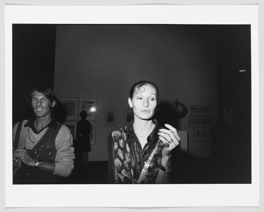 A woman at a gallery holds a cigarette and looks away while people stand in the background.