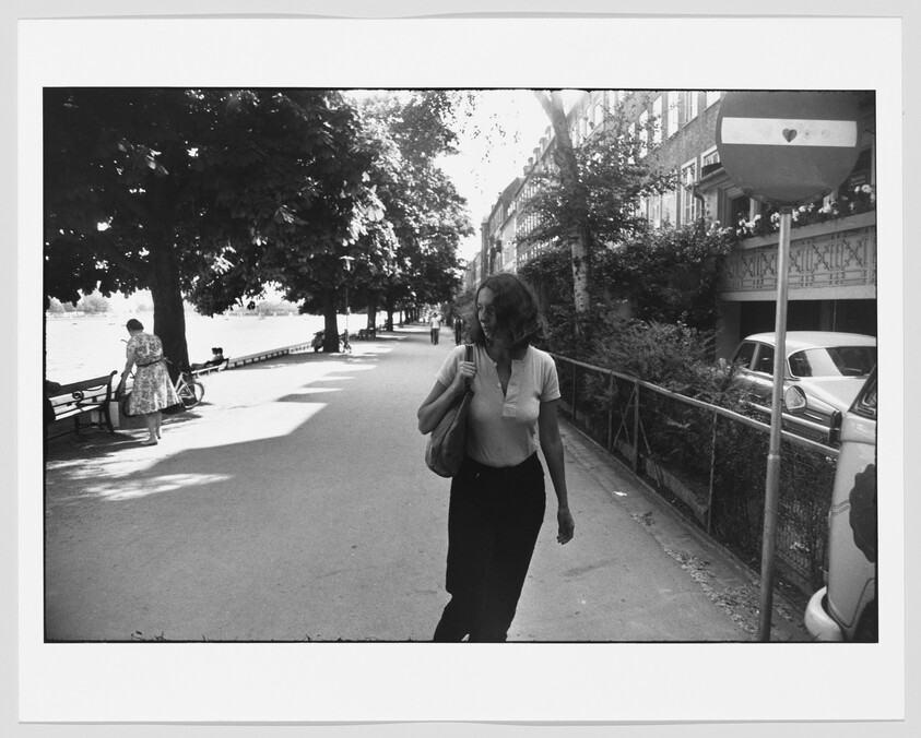 A woman walks along a tree-lined riverside path carrying a shoulder bag.